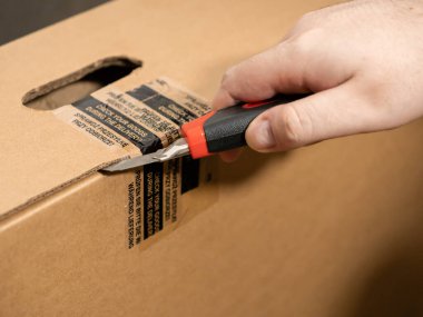 Mühürlü kahverengi karton nakliye kutusunu keskin bir bıçakla dikkatlice kesen kişinin yakın çekimi, tüketici güvenliği için güvenli teslimat ve paket denetimi vurgulanması
