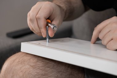Mobilyayı dikkatlice beyaz bir tahtaya sıkıştırmak için tornavida kullanan bir kişinin ellerinin yakın plan görüntüsü, gayretli ev yenileme veya pratik onarım görevlerini gösteriyor