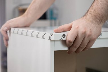 Yeni bir beyaz mobilya bileşeninin görünür geri dönüşüm sembolleriyle doğru bir şekilde hizalanmasını sağlayan bir adamın ellerine yakın çekim yapmak, modern bir ev kurma görevi sırasında dikkatli bir çaba gösterdiğini vurgulamak