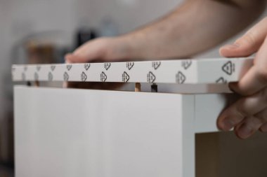 Kişinin ellerinin beyaz düz paket mobilya montajı, panelleri ahşap perdeler ve metal raptiyelerle tam olarak hizalama, pratik bir DIY görevi veya yeni bir ev düzeneği vurgulama ayrıntılı görünümü