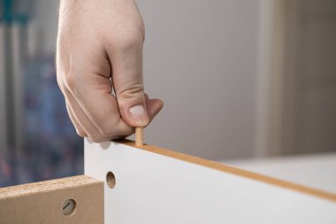 Yetişkin bir elin yakın plan çekimi ahşap bir iğneyi önceden delinmiş bir mobilya tahtasına yerleştirmek düz paket montajı ve DIY becerileri sergiliyor.