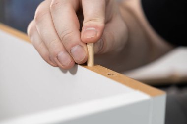 Yakın plan çekimde bir el, ustalıkla beyaz mobilya panelinin delinmiş deliğine ahşap bir panel iterek ev yapımı montajın hassasiyetini vurguluyor.