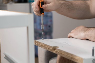 Beyaz parçacık tahtasına vida takmak için turuncu bir tornavida kullanarak ev gelişimini ve DIY mobilya montajını vurgulayan insan ellerinin ayrıntılı görüntüsü