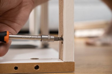 Bir DIY montaj sürecinde beyaz parçacık mobilyalarının bileşenlerini tutturmak için hassas bir tornavida kullanan bir kişinin elinin detaylı görüntüsü, kişisel çabayı ve ev yenilemesini temsil ediyor.