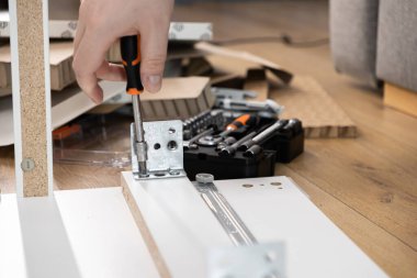 Elinde turuncu bir tornavida olan bir el beyaz mobilya tahtasına metal bir deste takıyor. Pratik DIY ve ev yenileme çalışmaları sergiliyor.