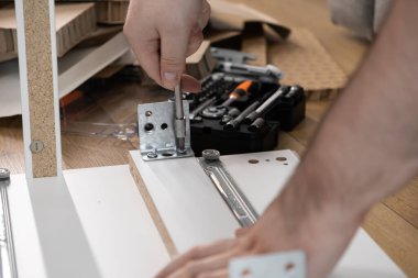 Mobilya montajı sırasında tornavidayla metal bir desteyi beyaz ahşap bir tahtaya bağlayan bir kişinin ellerinin yakın çekimi, ev yenilemesini veya kişisel bir projeyi vurguluyor