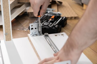 Beyaz mobilya tahtasındaki metal bir desteğe vida takmak için tornavidayı sıkıca kavrayan ellerin görüntüsü mobilya yapımı ve kişisel proje tamamlanmasının pratik sürecini vurguluyor