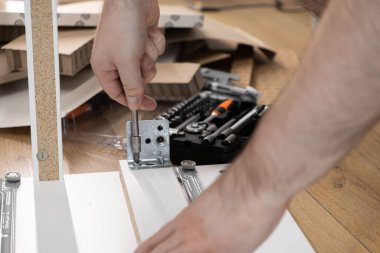 Bir insanın ellerinin yakın plan çekiminde tornavida kullanarak bir metal parçasını parçacık tahtası mobilyasının parçalarına iliştirirken, yanında bir alet kutusu ile ev yapımı DIY görevlerini vurguluyor.