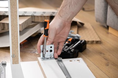 Yukarıdan aşağıya bakan bir görüntü, portakal kabzalı bir tornavida kullanarak başarılı bir ev montajı veya onarımını tasvir ederek mobilyaların üzerine metal bir deste yerleştiren bir erkek eli yakalar.