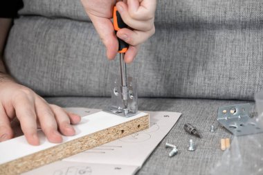 Tornavida kullanarak metal bir parçacığı parçacık tahtasına titizlikle yerleştiren ellerin yakın görüntüsü, adım adım mobilya montajını araçlarla ve kullanma kılavuzuyla gösteren