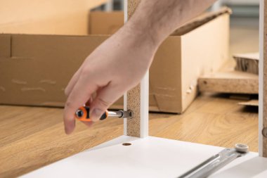 Turuncu bir tornavidayla yakın plan bir el beyaz parçacık tahtasına bağlanıyor, ev düzeneğini, DIY becerilerini ve yeni mobilya yapma sürecini temsil ediyor.