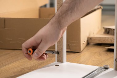 Yakın plan, yeni düz paket mobilya montajı, ev geliştirme, DIY çabaları ve modern mobilya kurulumu için tornavida kullanan bir kişinin elini gösteriyor.
