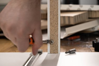 Bir kişinin elinin yakından görüntüsü, mobilyaları titizlikle monte ederken, beyaz parçacık paneline bir konnektör yerleştirmek için bir tornavida kullanarak, pratik ev yenilemesini vurguluyor