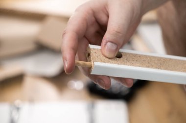 Beyaz mobilya panelinin delinmiş bir deliğine ahşap bir bağlantı panelini dikkatlice yerleştiren bir elin detaylı yakın plan görüntüsü, ev yapımı ve onarımı için kesin elle yapılan çalışmaların altını çiziyor