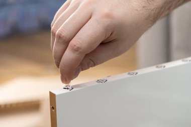 İnsan elinin beyaz parçacık tahtasından şeffaf koruyucu filmi titizlikle soyarken, talimatsal montaj rehberi sembollerini gösteren yakın plan görüntüler.