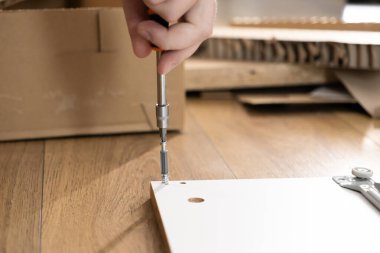 Bir insanın elinde bir tornavida tutarken detaylı bir fotoğraf, beyaz bir mobilyanın bileşenlerini titizlikle ahşap bir zemine yapıştırmak, ev yapımı montajı somutlaştırmak.