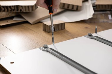 Bir kişinin eline bir tornavida ile dikkatlice beyaz bir mobilya paneline donanım iliştirerek pratik DIY becerilerini ve verimli ev düzenini gösteren bir yakın plan.