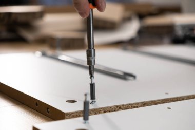 Beyaz parçacık tahtasına vida takmak için tornavida kullanan bir elin yakın plan çekimi, düz paket montajı, DIY projesi ve ev montaj çabalarını vurguluyor