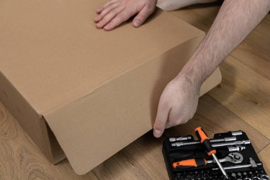 Bir adamın ellerini kapayarak düz kahverengi bir teslimat kutusunu açmaya başlarlar. Siyah bir alet çantası hazırdır. Ev dekorasyonu ya da mobilya montaj projesini betimler.