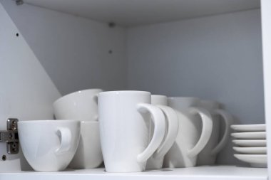 Beyaz seramik bardaklar ve tabaklar düzgün bir şekilde mutfak dolabının içindeki bir rafa yerleştirilmiş, verimli ev organizasyonunu ve aile için günlük temel kullanımını vurguluyor
