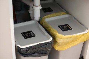 Mutfak tezgahının altındaki çöp kutularının yakın plan görüntüsü, uygun atık yönetimi ve sürdürülebilir yaşam uygulamaları için farklı etiketler içeriyor