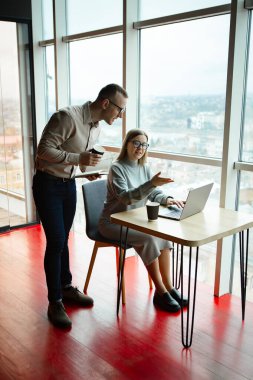 Ofiste iki çalışan var. Kadın dizüstü bilgisayarda çalışıyor, adam da arkasından monitöre bakıyor.