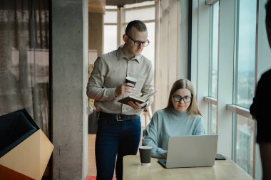 Ofiste iki çalışan var. Kadın dizüstü bilgisayarda çalışıyor, adam da arkasından monitöre bakıyor.