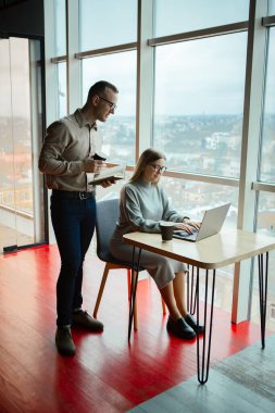 Ofiste iki çalışan var. Kadın dizüstü bilgisayarda çalışıyor, adam da arkasından monitöre bakıyor.