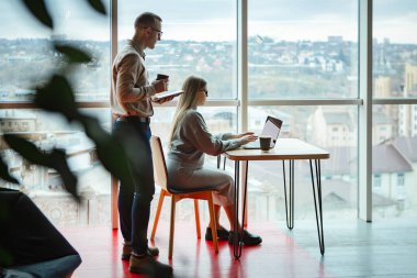 Ofiste iki çalışan var. Kadın dizüstü bilgisayarda çalışıyor, adam da arkasından monitöre bakıyor.
