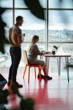 Ofiste iki çalışan var. Kadın dizüstü bilgisayarda çalışıyor, adam da arkasından monitöre bakıyor.