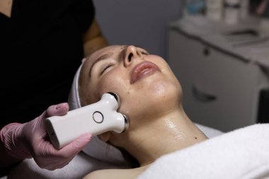 Mikro akım cihazıyla estetik yüz tedavisi. Pembe eldivenli estetisyen çene hattı boyunca masaj yapıyor. Güzellik kliniğinde profesyonel cilt bakımı.