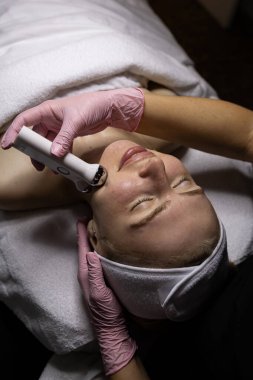 Mikro akım cihazıyla estetik yüz tedavisi. Pembe eldivenli estetisyen çene hattı boyunca masaj yapıyor. Güzellik kliniğinde profesyonel cilt bakımı.