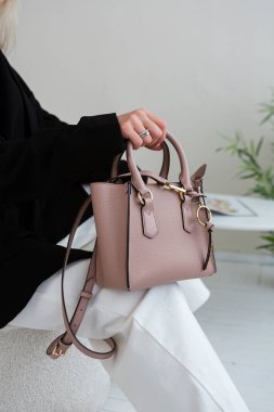 Üzerinde altın rengi detaylar olan küçük kahverengi deri bir çanta. Yapılandırılmış tasarım. Siyah ceket ve beyaz pantolon.