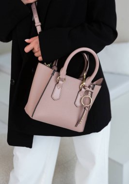 Üzerinde altın rengi detaylar olan küçük kahverengi deri bir çanta. Yapılandırılmış tasarım. Siyah ceket ve beyaz pantolon.