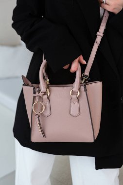 Üzerinde altın rengi detaylar olan küçük kahverengi deri bir çanta. Yapılandırılmış tasarım. Siyah ceket ve beyaz pantolon.