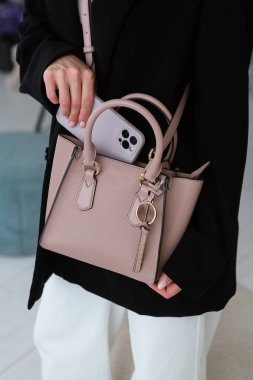 Üzerinde altın detaylar olan küçük, kahverengi deri bir çanta, yapılandırılmış tasarım, siyah ceket ve beyaz pantolon, içinde telefon..