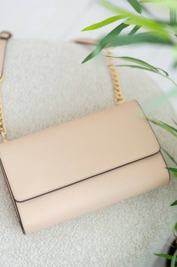 Altın zincir kayışlı bej deri bir çapraz ceset torbası, minimalist tasarım, yeşil bitki detayı ile dokulu kumaş üzerine yerleştirilmiş..