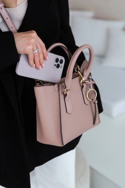 Üzerinde altın detaylar olan küçük, kahverengi deri bir çanta, yapılandırılmış tasarım, siyah ceket ve beyaz pantolon, içinde telefon..