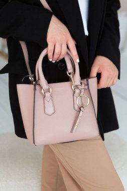 Üzerinde altın rengi detaylar olan küçük kahverengi deri bir çanta. Yapılandırılmış tasarım. Siyah ceket ve beyaz pantolon.