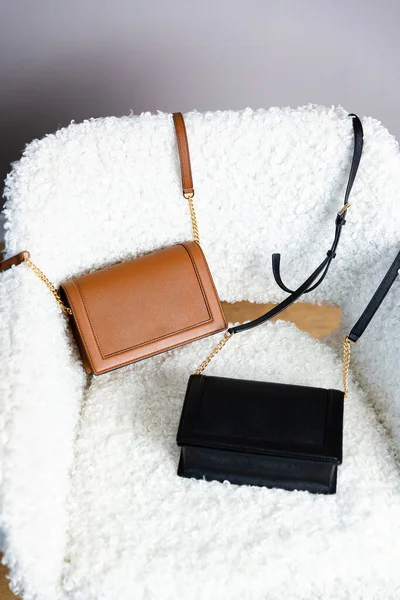 Sandalyede iki el çantası var. Siyah ve kahverengi kadın çantaları açık minimalizmin içinde beyaz sandalyede sergilenmektedir.