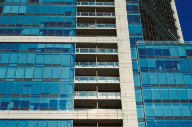 Bir kentsel alanda yüksek bir bina duruyor. Cam pencereleri güneş ışığını yansıtır. Balkonlar birçok kattan görülebilir. Gökyüzü açık ve mavi.