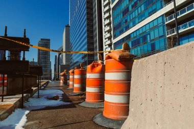 Trafik konileri, gündüz açık mavi gökyüzünün altındaki yüksek binaların yanındaki alanı güvenlik şeridiyle bir inşaat alanı boyunca yerleştiriliyor..