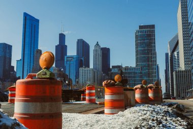 İşçiler bir kentteki bir inşaat alanında mevcuttur. Turuncu koniler bölgeyi işaretliyor. Kışın açık mavi bir gökyüzünün altında bölgenin etrafını saran yüksek binalar. Kar zeminin bazı kısımlarını kaplıyor..