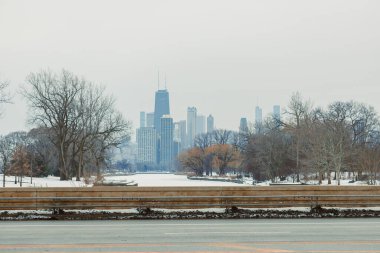 Chicago 'nun ufuk çizgisi manzaralı soğuk kış günü. Kar zemini kaplıyor ve ağaçlar çıplak. Ön planda, görünürde araba olmayan sessiz bir atmosfer gösteren bir yol var..