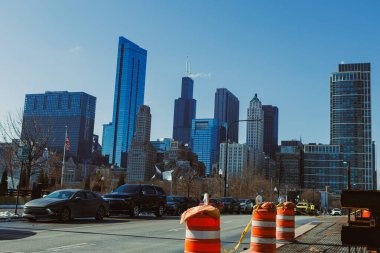 Arkasında yüksek binalar olan bir şehir silueti manzarası. İnşaat bariyerleri cadde boyunca görülüyor. Arabalar güneşli bir günde yakınlarda park edilir..