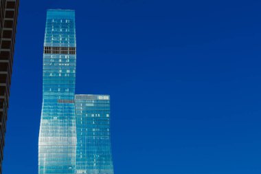 Yoğun bir şehirde, uzun cam binalar parlak mavi gökyüzüne doğru uzanır. Yapılar güneş ışığını yansıtıyor ve aşağıdaki kentsel yaşamın etkinliğini gösteriyor..