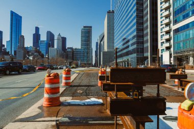 Şehir caddesinde birkaç trafik engeli ve inşaat malzemesi kuruldu. Mavi gökyüzünün altında iki taraf da yüksek binalar var. Araçlar yakındaki yolda görülebiliyor..