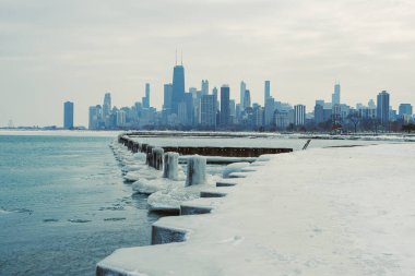 Chicago ufuk çizgisi kışın göl kıyısı boyunca buz ve karlı yüksek binalara sahiptir. Sahne, üzerinde gri bulutlar ve kıyıda donmuş su olan soğuk bir günü gösteriyor..