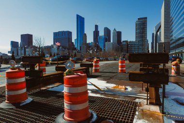 İşçiler mavi gökyüzünün altındaki yüksek binaların önündeki turuncu ve beyaz bariyerleri kullanarak inşaat işleriyle meşguller. Araçlar yakınlarda park halinde..
