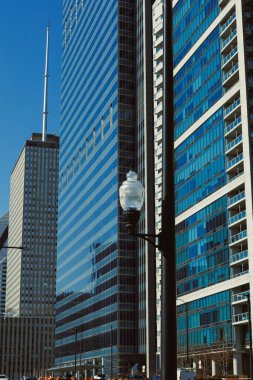 Şehir merkezindeki uzun binalar açık bir günde güneş ışığını yansıtır. Sokak lambaları kaldırım boyunca işlek yolun yanında durur. Sahne modern mimariyi gösteriyor..